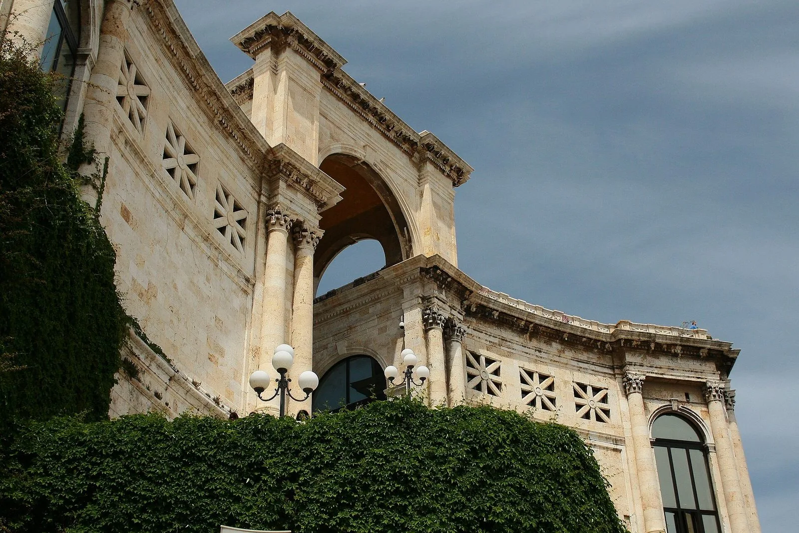 Il Bastione di Saint Remy a Cagliari visto dal basso: facciata in pietra chiara con colonne, arco monumentale e lampioni, edera verde in primo piano e cielo blu