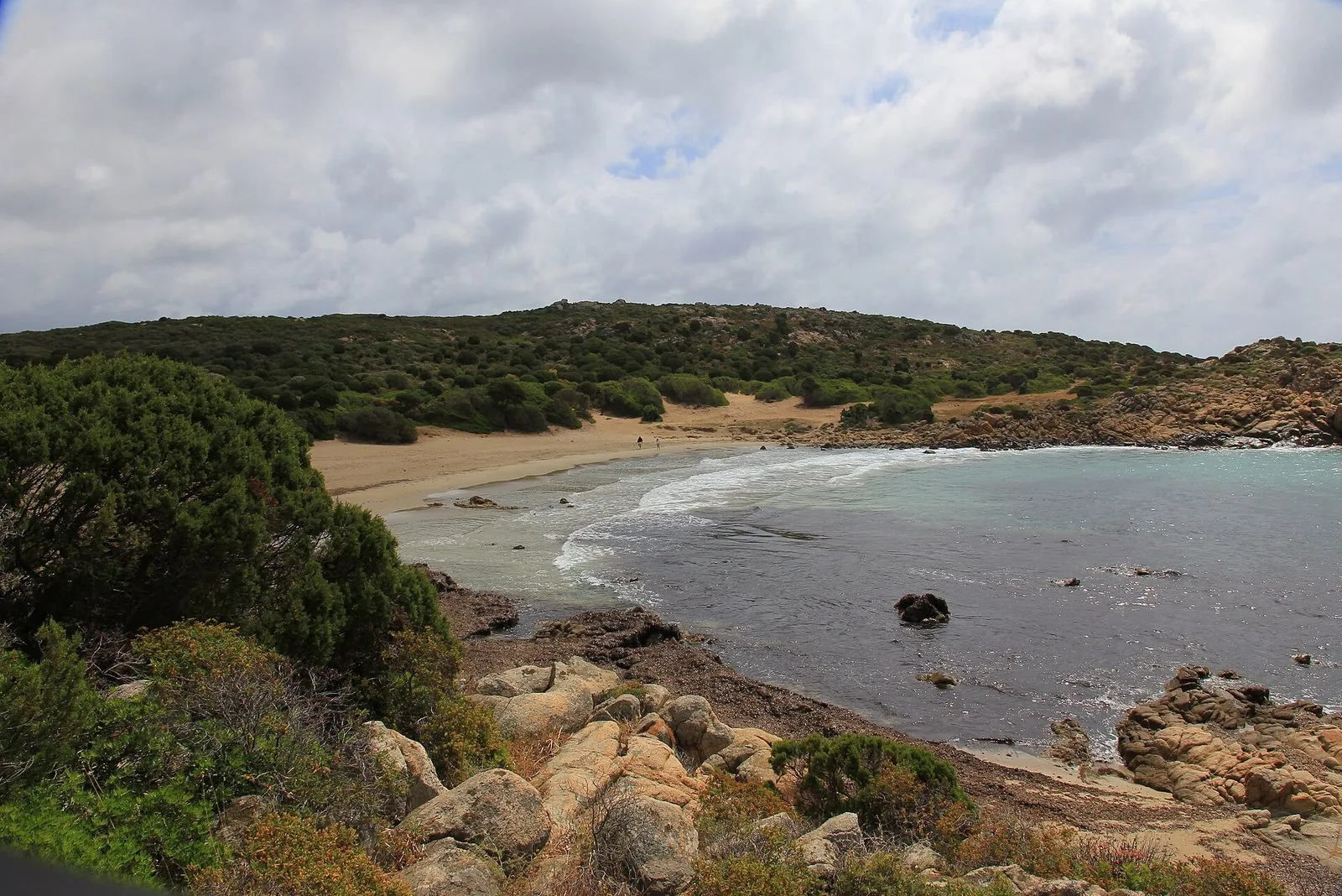 Cala Cipolla sotto un cielo nuvoloso: spiaggia di sabbia chiara, onde leggere, rocce granitiche e colline coperte di macchia mediterranea