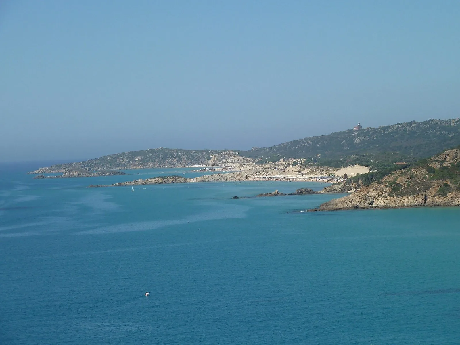 La costa di Chia vista dall'alto: mare turchese, un promontorio roccioso con macchia mediterranea e, in lontananza, la spiaggia e il faro di Capo Spartivento