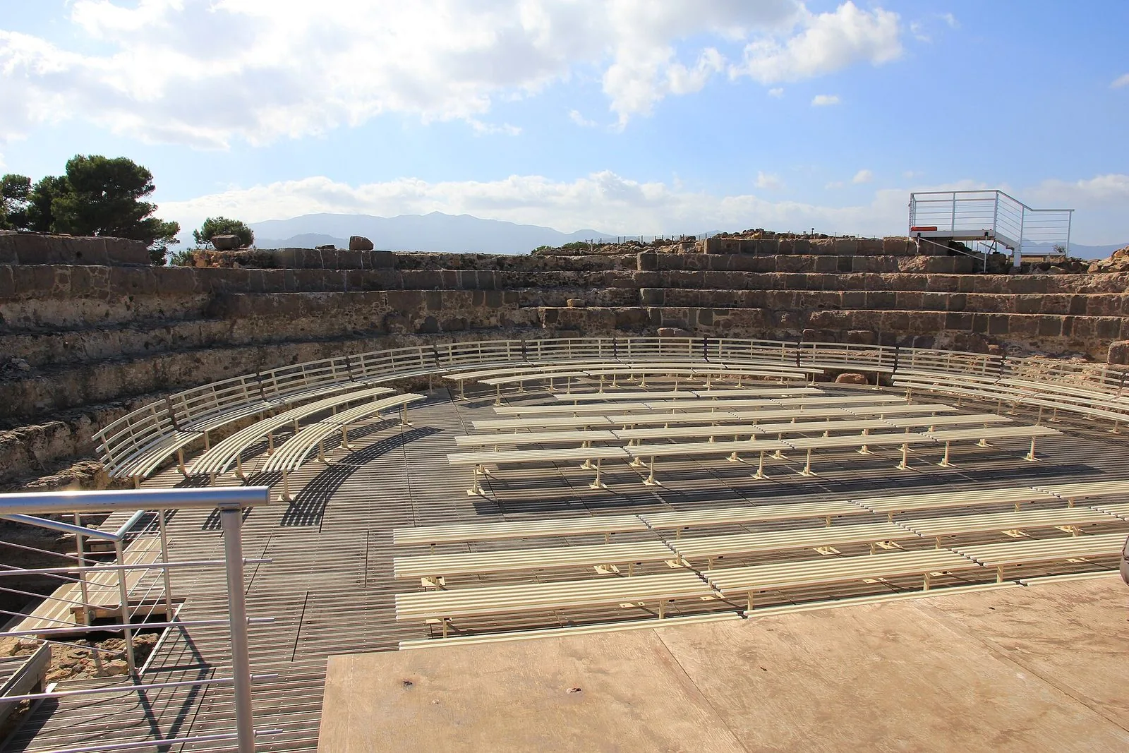La cavea in pietra del teatro romano di Nora con le panche chiare allestite per gli spettacoli, sullo sfondo le montagne e un cielo azzurro con nuvole