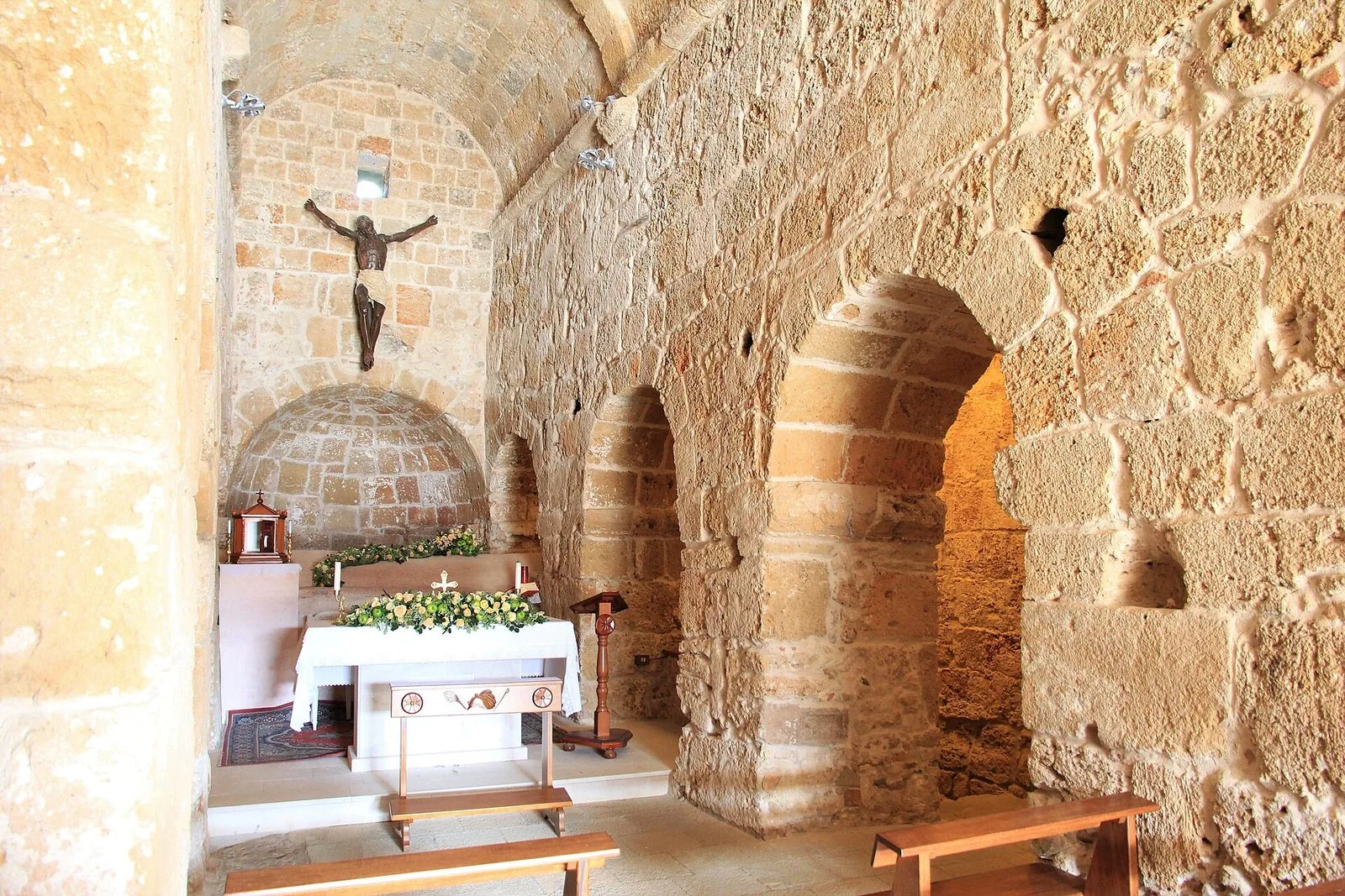 L'interno in pietra chiara della chiesa di Sant'Efisio a Nora, con l'altare ornato di fiori, il crocifisso appeso alla parete di fondo e le arcate laterali