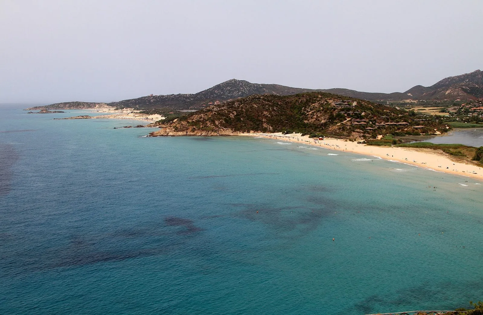 Vista dall'alto della baia di Chia: la spiaggia di sabbia chiara, il mare turchese, il promontorio coperto di macchia e, sullo sfondo, le altre spiagge fino a Capo Spartivento