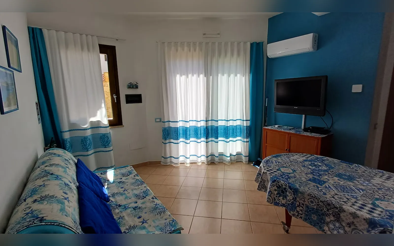 Il soggiorno con il divano letto azzurro, la parete blu con la TV, il climatizzatore e le tende bianche e azzurre